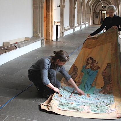 Monika von Bernuth (l.) und Vera Sous halten ein großes Stück Stoff, auf dem Ghada Khano als heiliger Christopherus dargestellt wird. Sie befinden sich im Kreuzgang von Kloster Steinfeld.