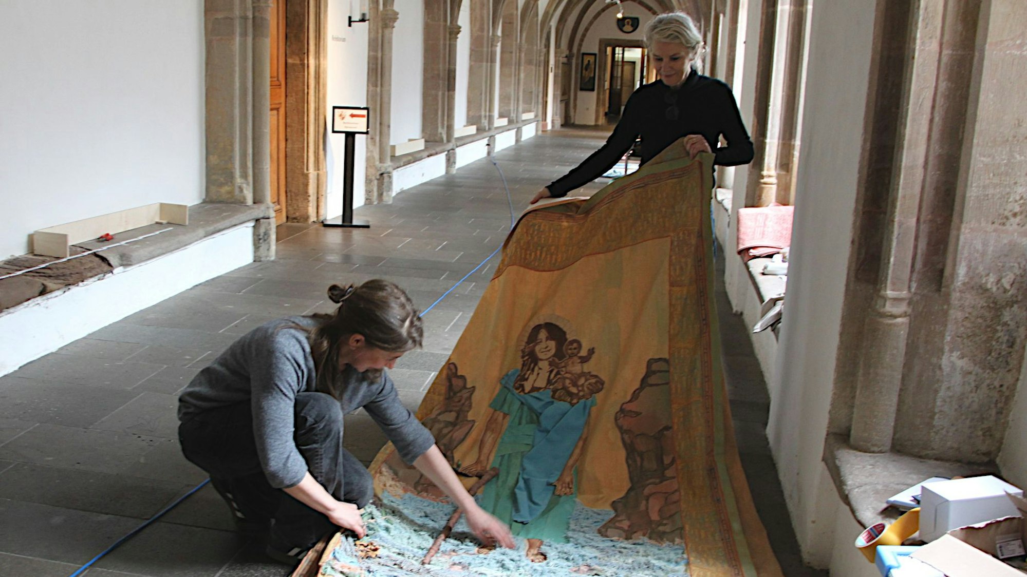 Monika von Bernuth (l.) und Vera Sous halten ein großes Stück Stoff, auf dem Ghada Khano als heiliger Christopherus dargestellt wird. Sie befinden sich im Kreuzgang von Kloster Steinfeld.