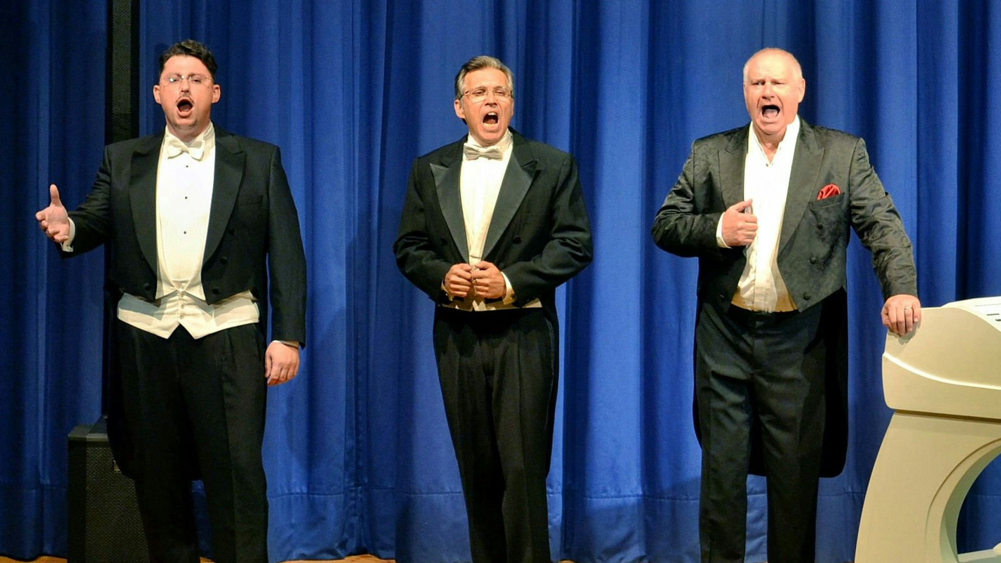 Die Tenöre Johannes Groß, Oscar Marin und Ricardo Marinellostehen singen in Anzügen auf der Bühne des Euskirchener Stadttheaters. Neben ihnen spielt Claudia Hirschfeld eine elektrische Orgel.