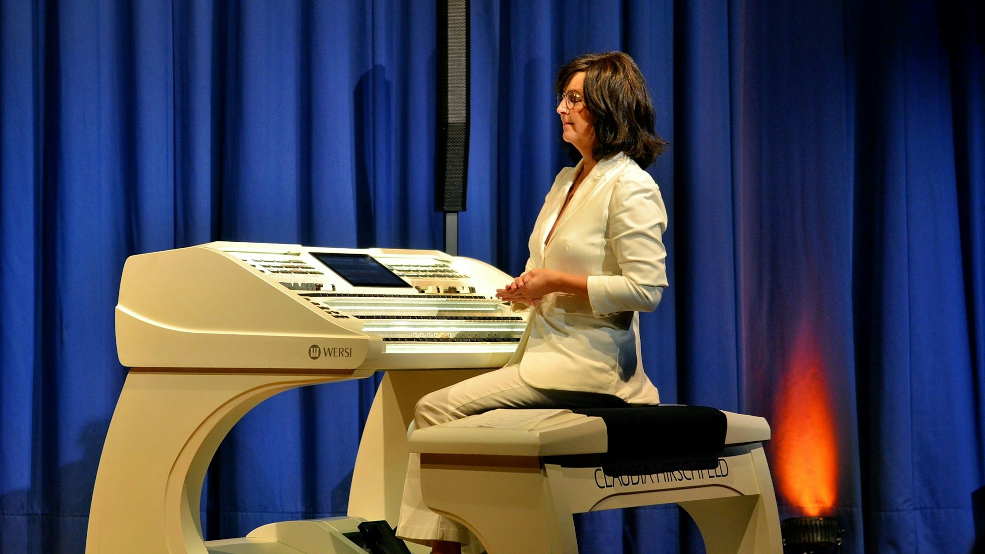 Claudia Hirschfeld sitzt mit gefalteten Händen an einer elektronischen Orgel. Im Hintergrund befindet sich ein Bühnenvorhang.