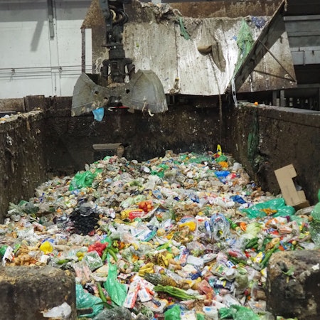 Blick auf den Inhalt eine Betonwanne: Ein geöffneter Baggergreifer schwebt über Verpackungen und Lebensmitteln.