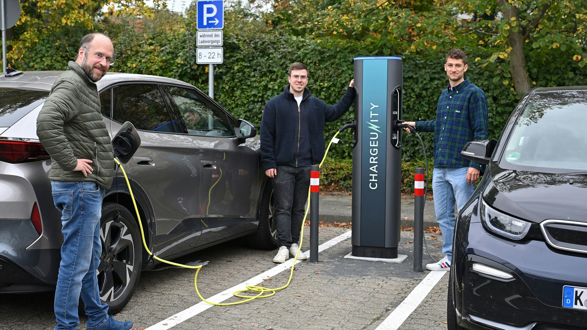 Drei Männer stehen an einer E-Ladesäule. Daneben zwei Autos