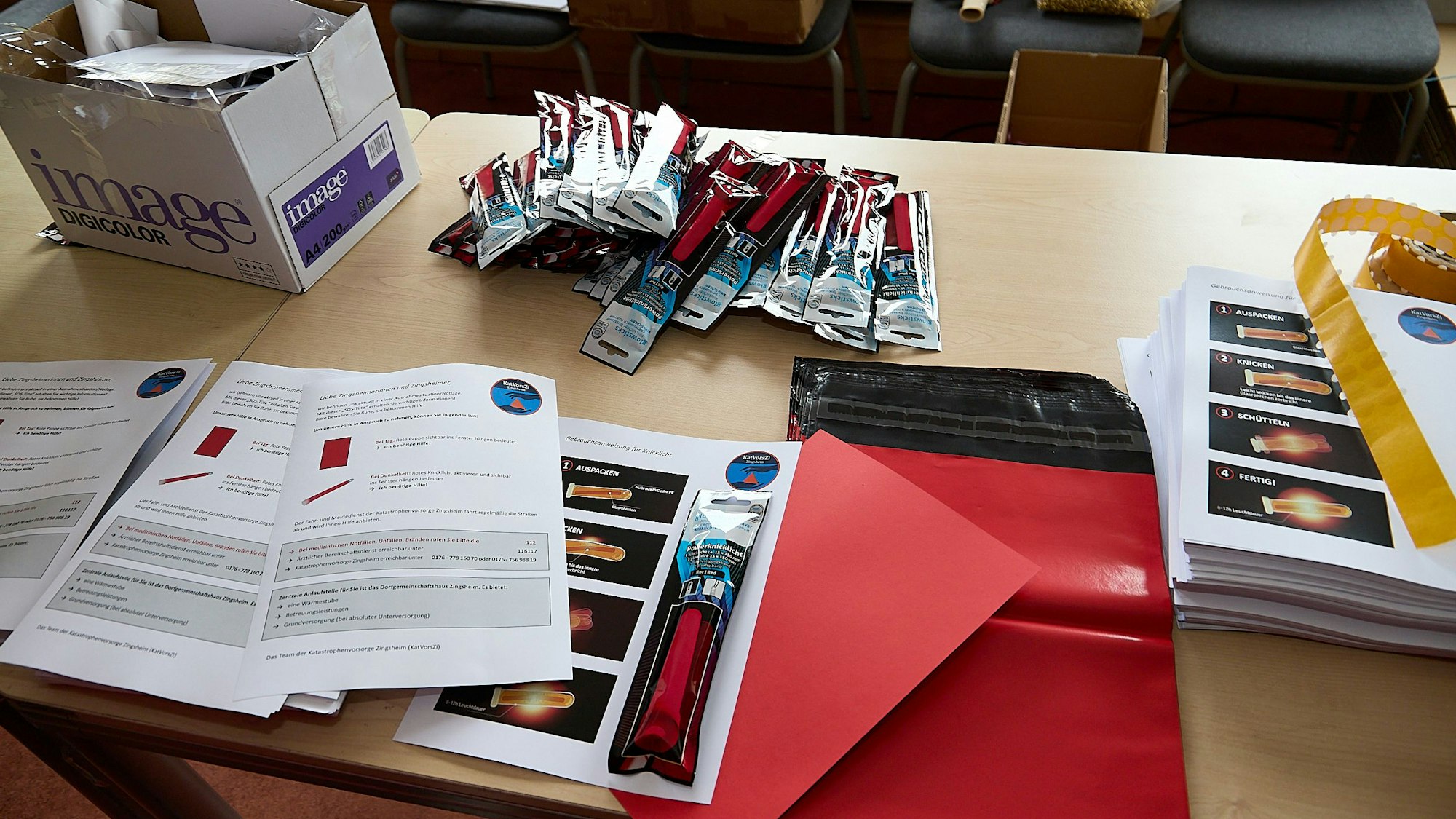Eine rote Pappe, ein Knicklicht und ein analoges Tutorial sind in der SOS-Tüte enthalten. Die Materialien liegen auf einem Tisch.