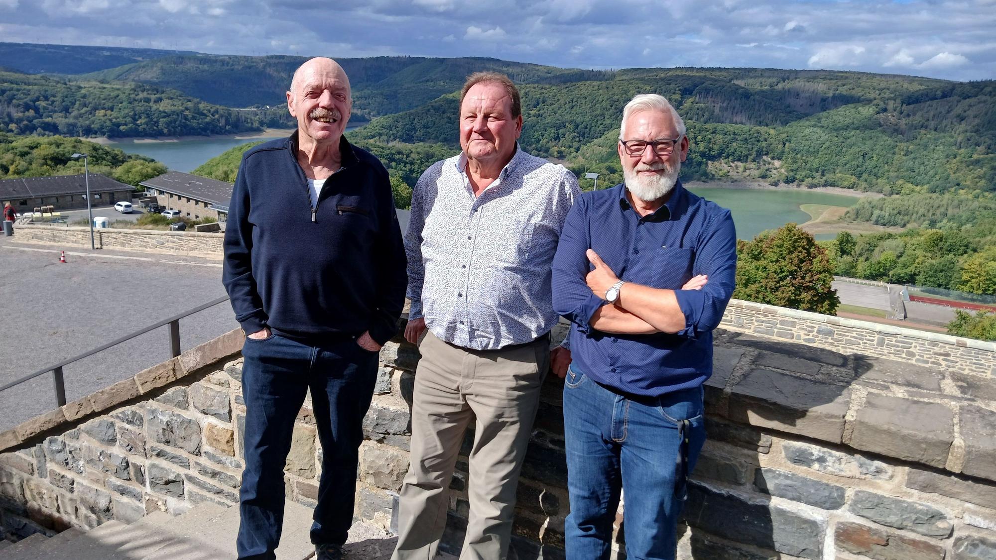 René Claeys (v.l.), Luc Bruylandt und Jean-Marie Malaise stehen in Vogelsang vor einer Mauer. Im Hintergrund ist der Urftsee in einer grünen Landschaft zu sehen.