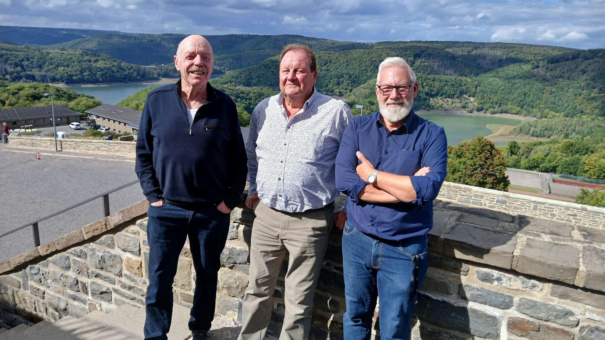 René Claeys (v.l.), Luc Bruylandt und Jean-Marie Malaise stehen in Vogelsang vor einer Mauer. Im Hintergrund ist der Urftsee in einer grünen Landschaft zu sehen.
