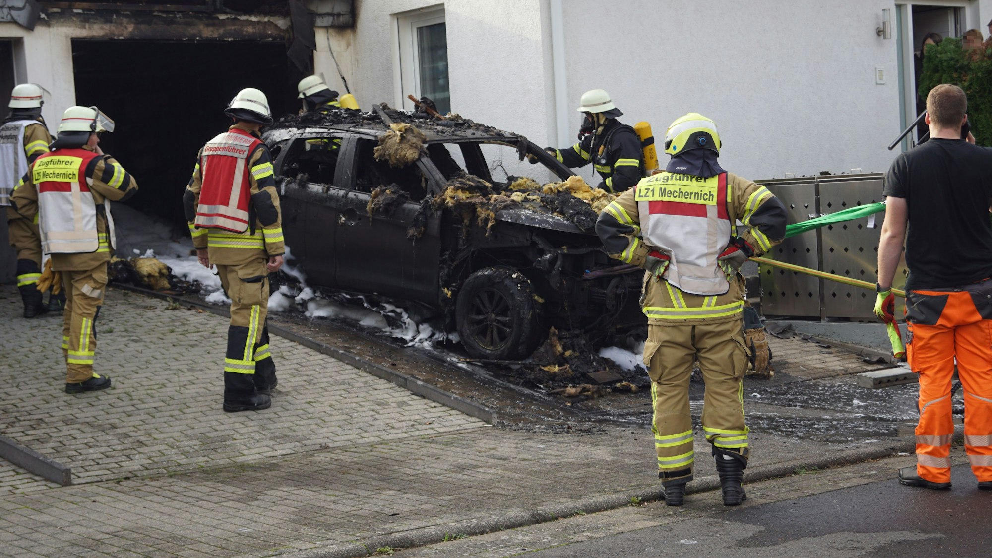 Das Bild zeigt den ausgebrannten Wagen vor der Garage. Einige Feuerwehrleute betrachten es.