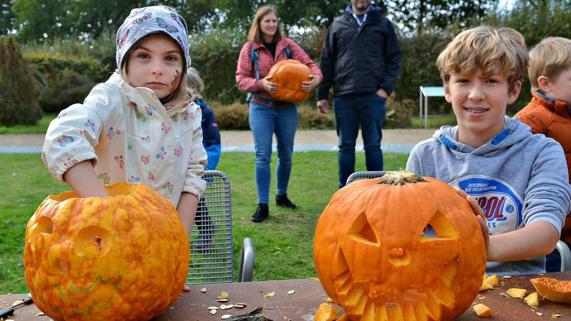 Ein Mädchen greift in das Innere eines Kürbisses, in den eine Fratze geschnitzt wurde. Neben ihr sitzt ein Junge, der seine Hand auf einen weiteren Kürbis mit Fratze gelegt hat.