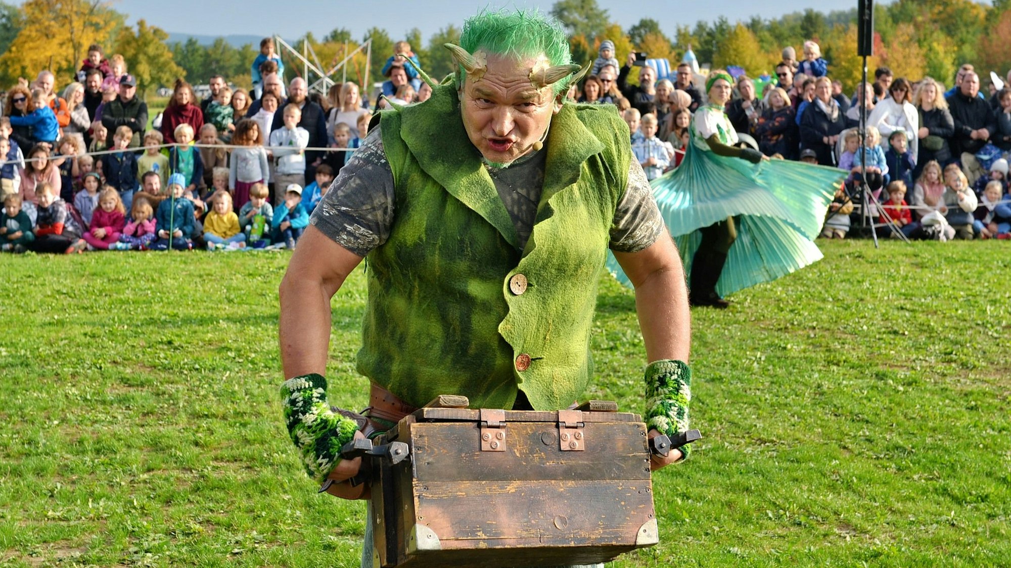 Ein Mann mit grün gefärbten Haaren, aufgesetzten Hörnern und einer Weste trägt eine Truhe. Im Hintergrund sitzen Kinder und Erwachsene und schauen ihm zu.
