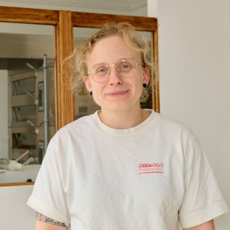 Ein Frau mit blondem Haar und Brille steht vor einer Scheibe, dahinter ist die Backstube zu sehen, in der sie Brot und Croissants backt.