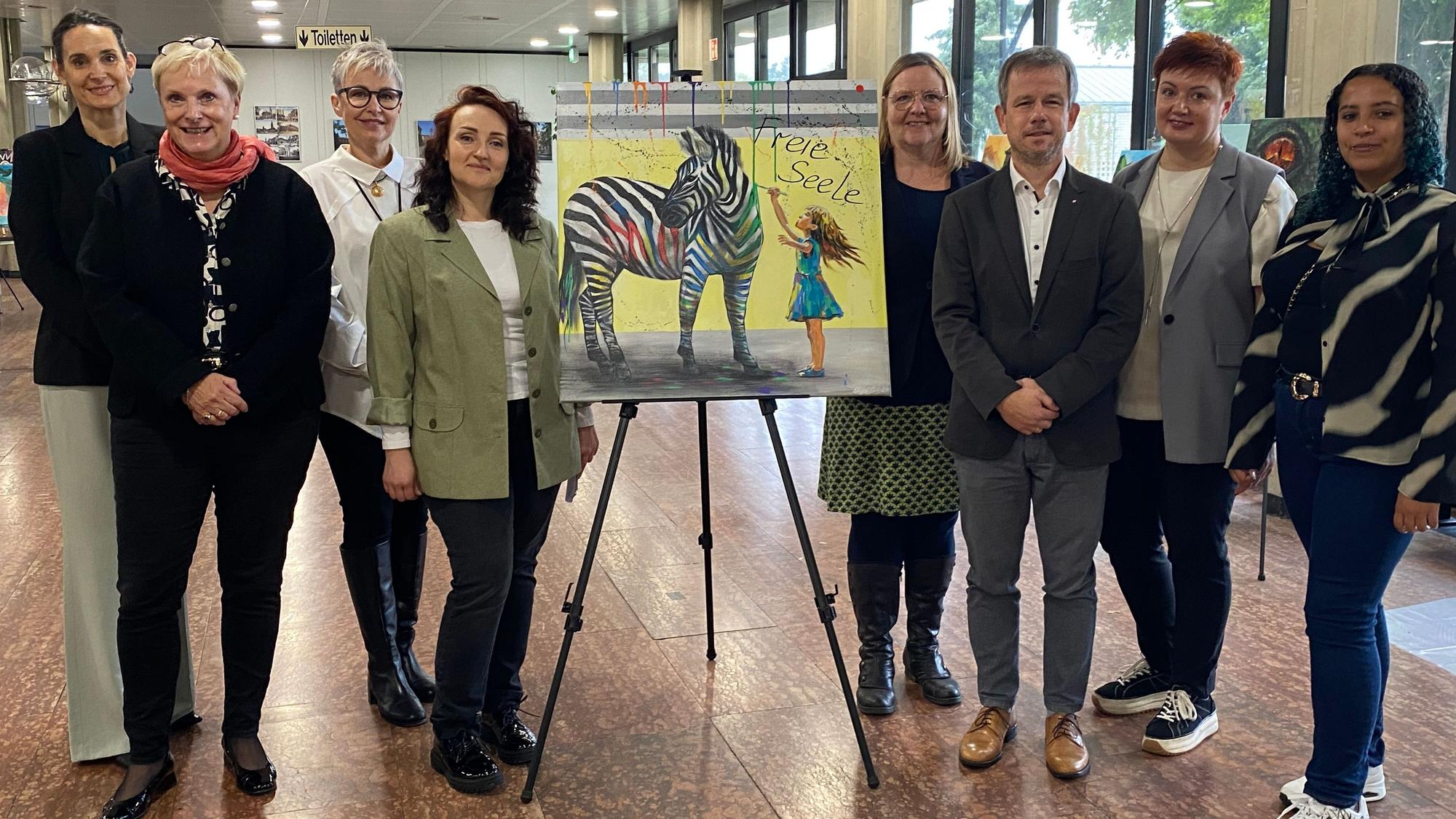 Vertreter des Kreises Euskirchen und des Caritasverbandes Euskirchen versammeln sich um das Talisman-Bild „Freie Seele“, auf dem ein Mädchen zu sehen ist, das ein Zebra mit bunten Streifen verziert.