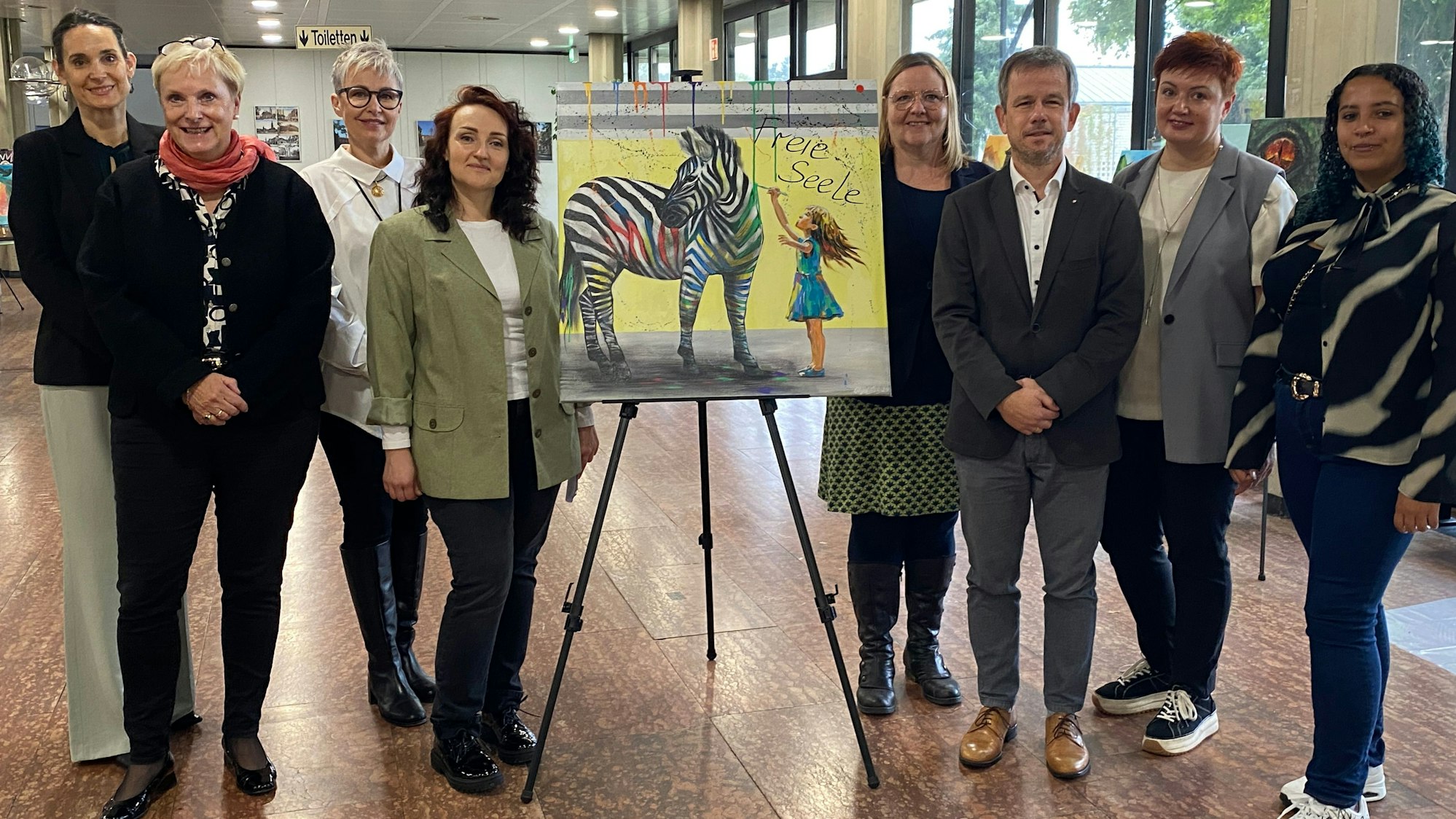 Vertreter des Kreises Euskirchen und des Caritasverbandes Euskirchen versammeln sich um das Talisman-Bild „Freie Seele“, auf dem ein Mädchen zu sehen ist, das ein Zebra mit bunten Streifen verziert.