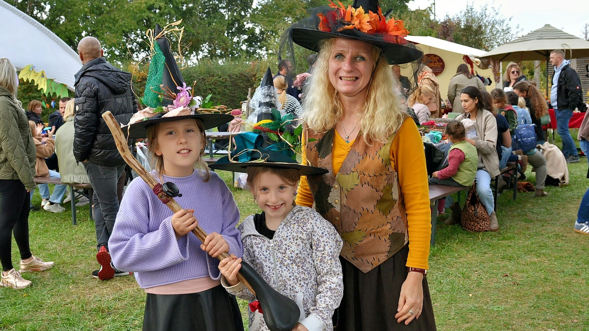 Zwei Mädchen tragen Hexenmützen und halten gemeinsam einen Besen. Neben ihnen steht eine Frau, die ebenfalls als Hexe verkleidet ist.