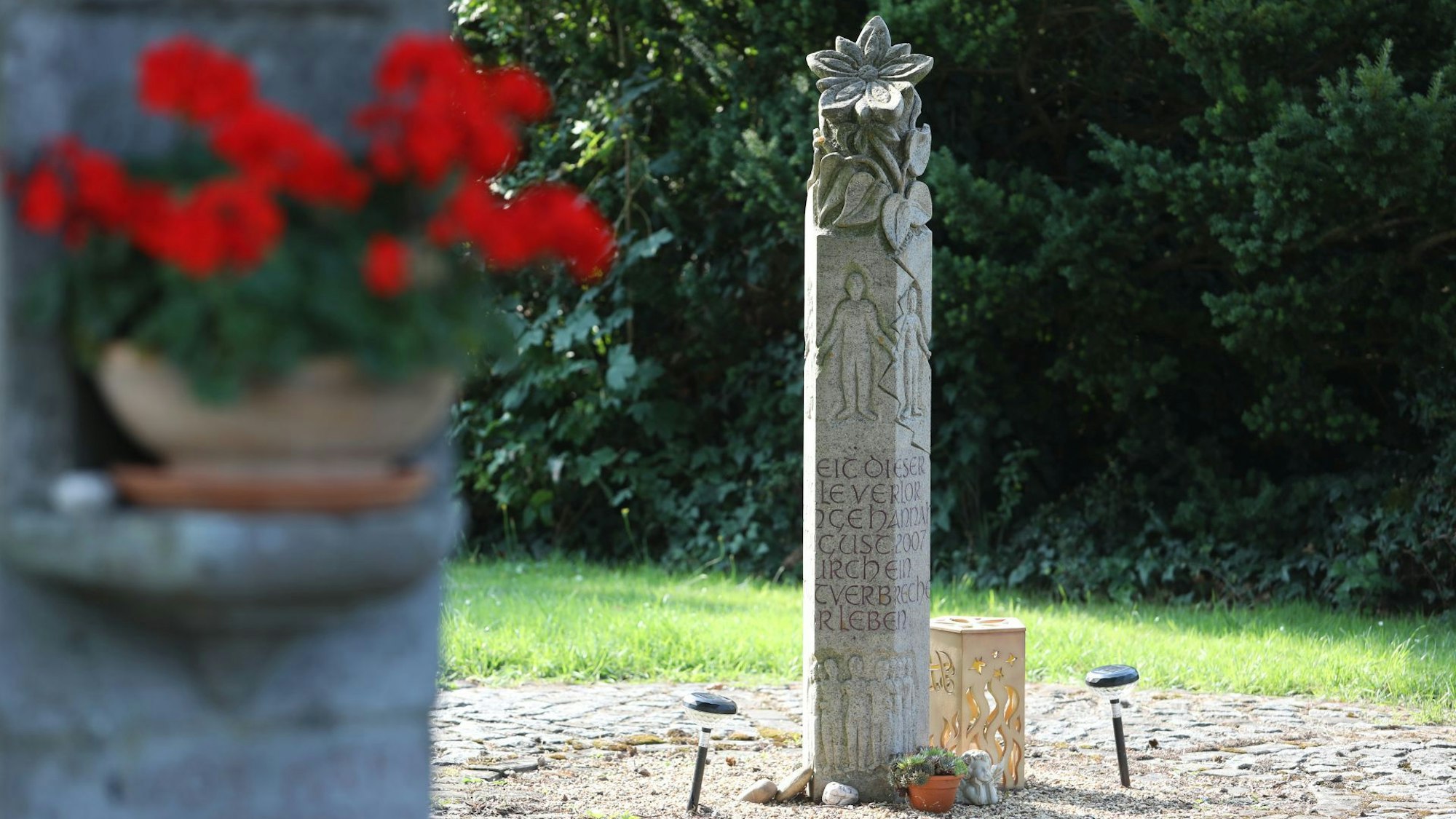 Im Vordergrund ein Blumtentopf mit roten Blüten, im Hintergrund ein Gedenkstein.