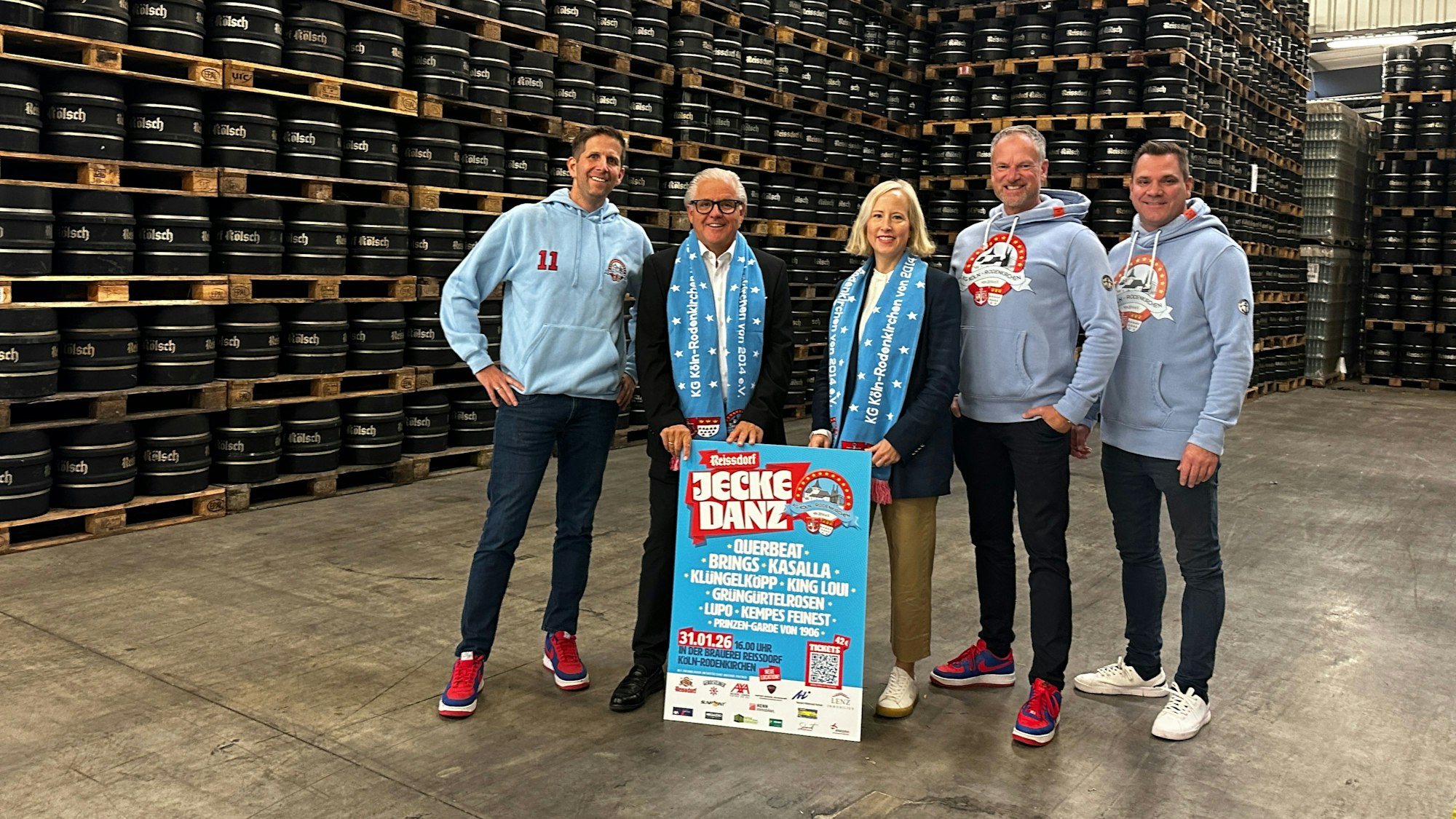 In einer großen Lagerhalle mit unter die Decke gestapelten Kölschfässern stehen vier Männer und eine Frau.