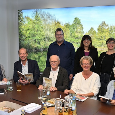 Mehrere Frauen und Männer sitzen an einem Tisch und halten ein Buch in der hand. Hinter ihnen eine grüne Landschaft.