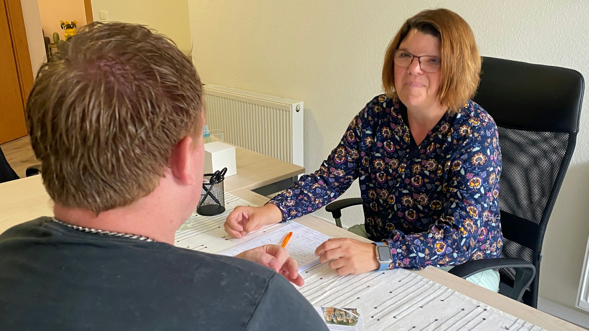 Katja Eichelbaum sitzt am Schreibtisch und schaut ihren Klienten an, der von hinten zu sehen ist.