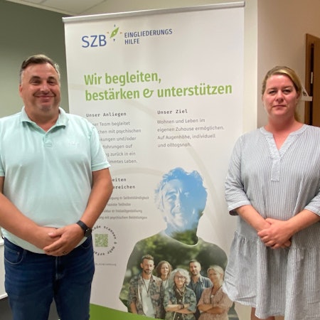 Thomas Sohr und Jessica Breitbarth stehen vor einem Banner, auf dem die SZB für ihre Angebote wirbt.