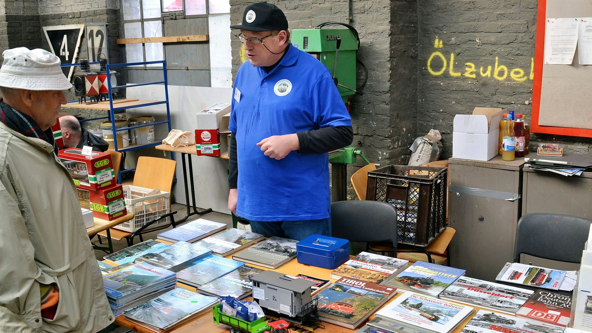 Zwei Männer stehen an einem Tisch, auf dem zahlreiche Bücher liegen, und unterhalten sich.