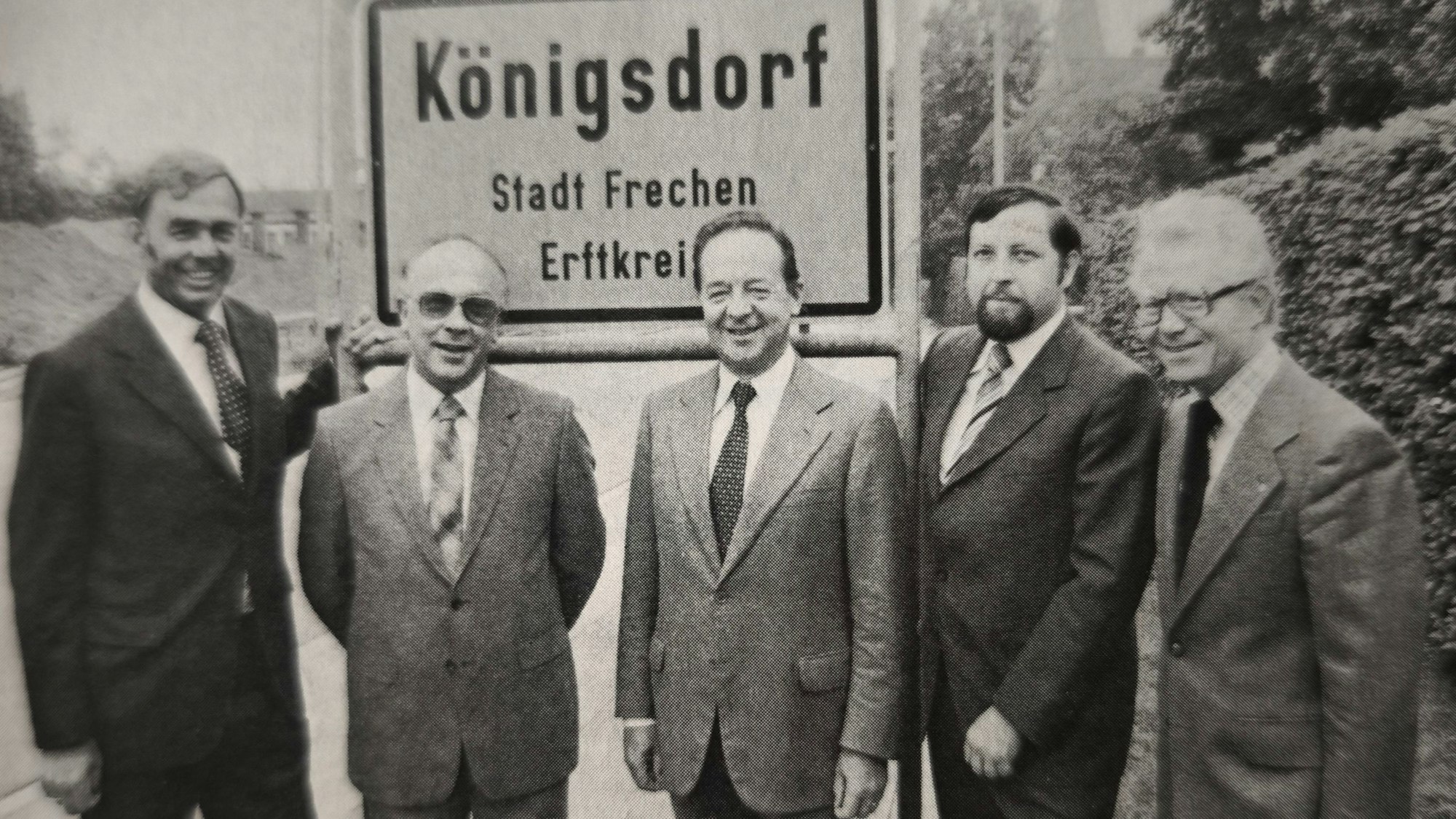 Das Bild ist schwarz-weiß und zeigt die fünf Herren im Anzug vor dem Ortseingangsschild zu Königsdorf.