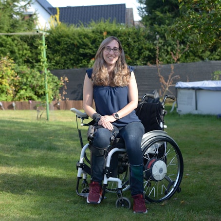 Die Autorin sitzt in einem Garten im Rollstuhl und lächelt in die Kamera.