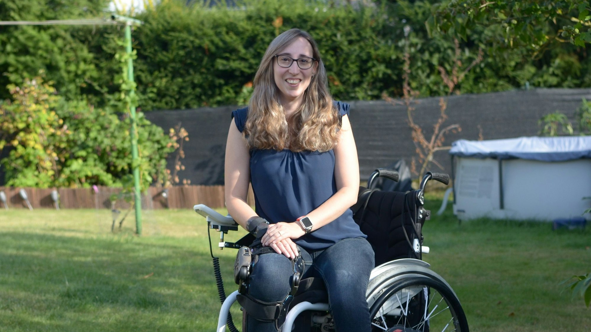 Die Autorin sitzt in einem Garten im Rollstuhl und lächelt in die Kamera.