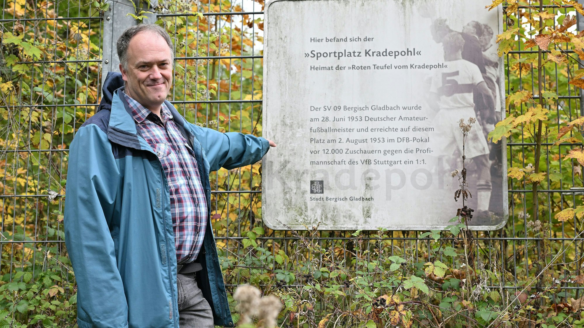 Ein Mann mit blauer Windjacke steht vor einem Gitter mit einem Schild. Darauf steht: „Hier befand sich der Sportplatz Kradepohl. Heimat der RotenTeufel.“