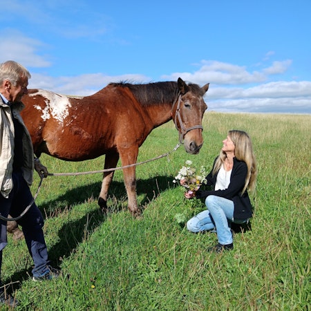 Eine Frau hält einen Blumenstrauß in der Hand und kniet auf einer Wiese vor einem braunen Pferd. Ein Mann hält das Pferd an einem Strick.