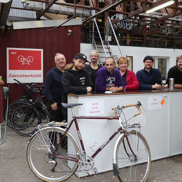 Mehrere Menschen stehen hinter einer Art Theke, daran lehnt ein Fahrrad, weitere Kinderräder stehen am Rand.