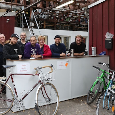 Mehrere Menschen stehen hinter einer Art Theke, daran lehnt ein Fahrrad, weitere Kinderräder stehen am Rand.