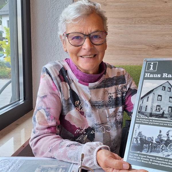 Renate Krumpen sitzt an einem Tisch und hält eine Tafel in der Hand, die alte Ansichten eines Hauses zeigt.