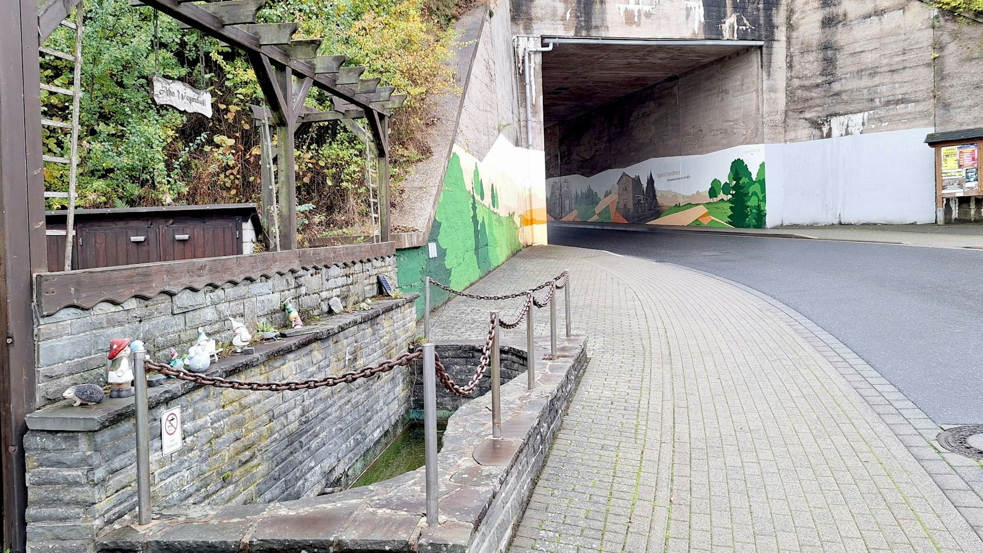 Vor der mit Graffiti versehenen Bahnunterführung in Dahlem-Schmidtheim ist links ein Wasserbecken, das mit einer Kette gesichert ist.