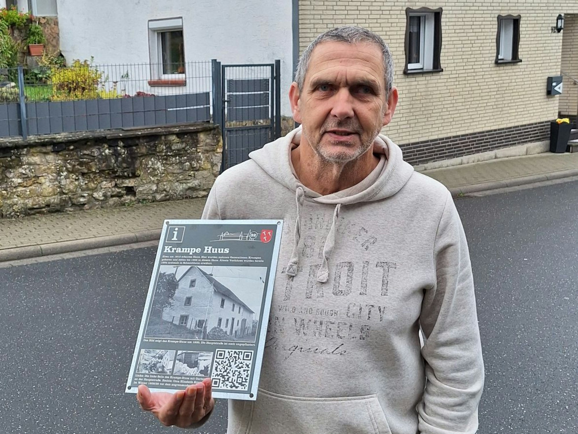 Ein Mann steht in Dahlem-Schmidtheim auf einer Straße vor einem Haus. Er hält eine Tafel mit alten Ansichten des Hauses in der Hand.