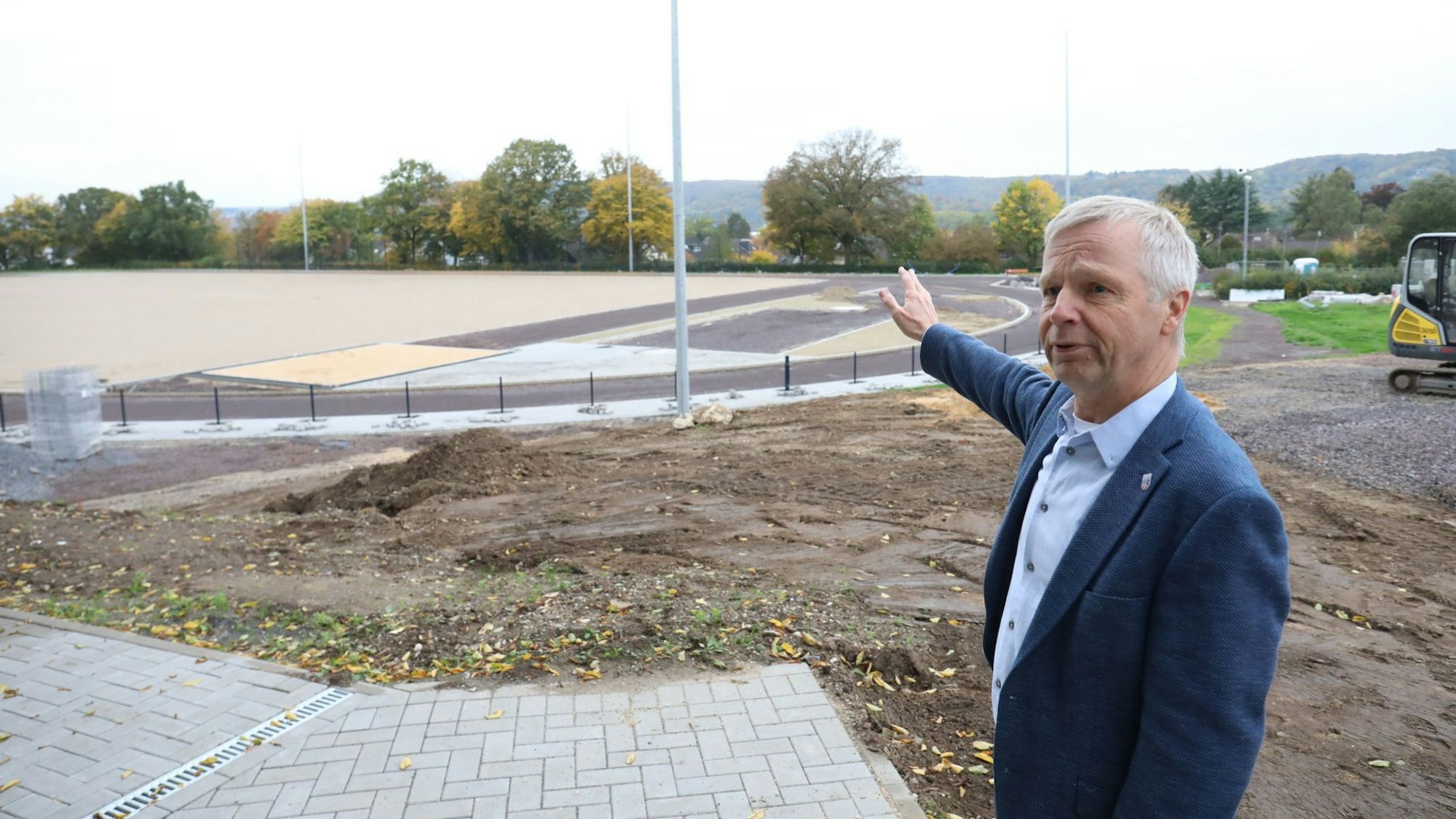Ein Mann im blauen Sakko am Rande einer Baustelle, im Hintergrund das Stadion am Menzenberg, das saniert wird.