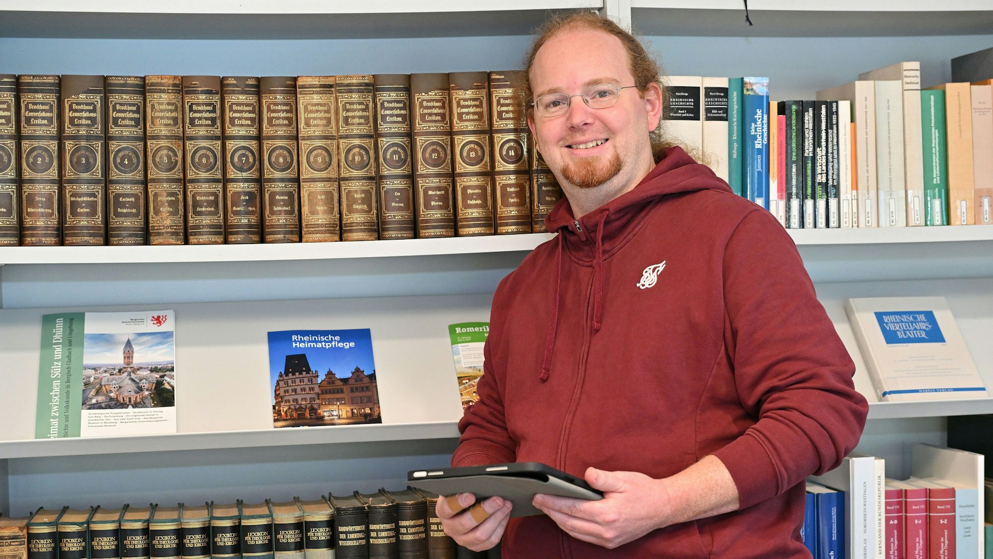 Ein Mann in rotem Kapuzenpulli, mit rotem Haar und rotem Bart, steht vor einem Bücherregal mit alten Nachschlagewerken.