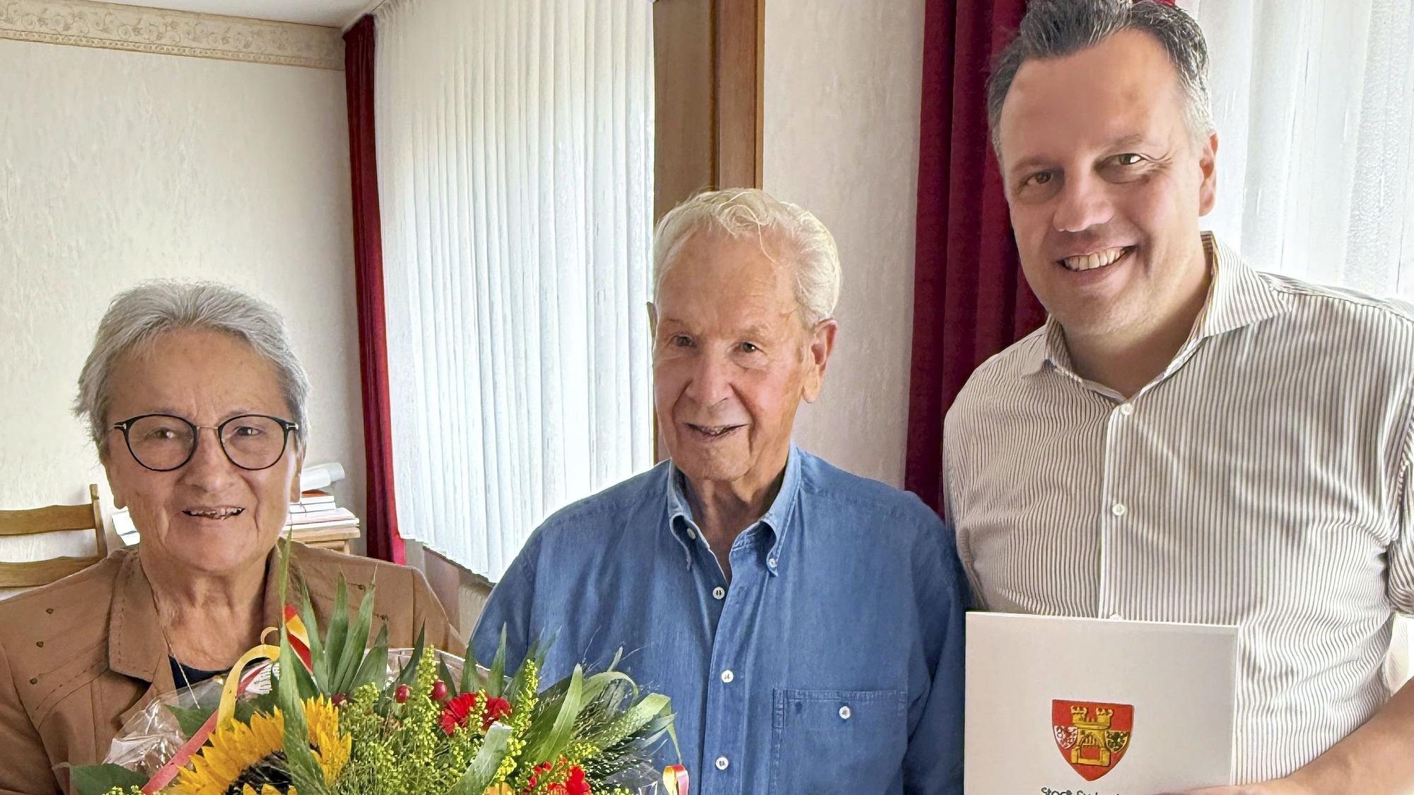 Anneliese Büschkes hält einen Blumenstrauß, Bürgermeister Sacha Reichelt überreicht Hans-Dieter Büschkes eine Urkunde, auf der das Wappen der Stadt Euskirchen zu sehen ist.