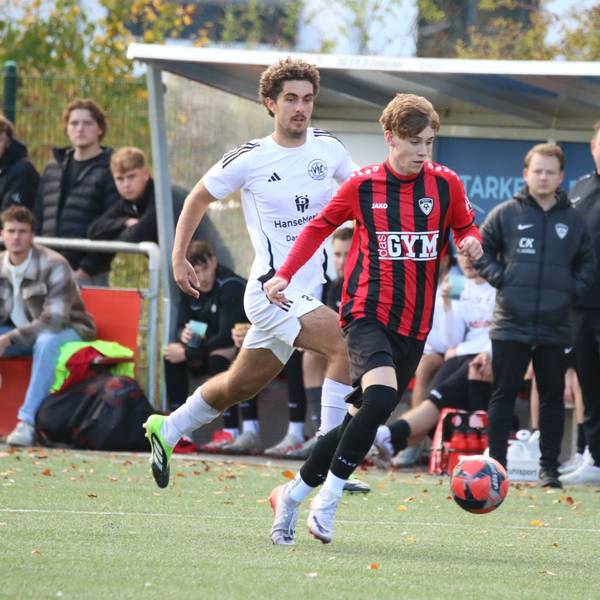 Aaron Mertens läuft mit dem Ball, ein Gegenspieler verfolgt ihn. Im Hintergrund gucken Trainer und Betreuer der JSG Erft 01 zu.