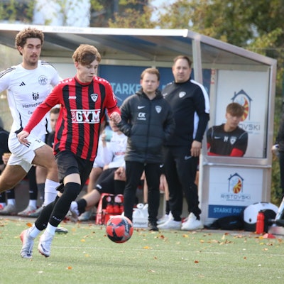 Aaron Mertens läuft mit dem Ball, ein Gegenspieler verfolgt ihn. Im Hintergrund gucken Trainer und Betreuer der JSG Erft 01 zu.