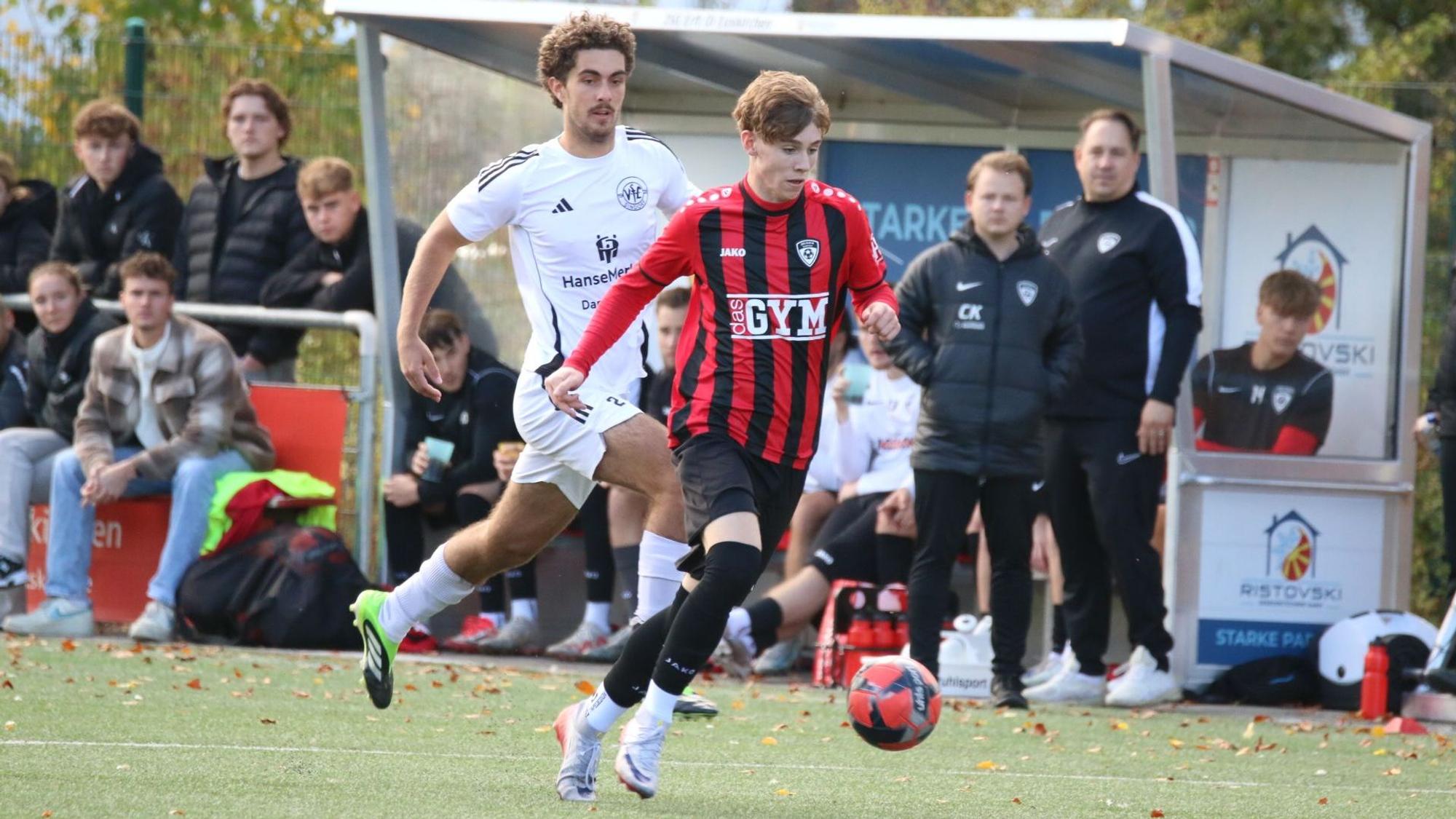 Aaron Mertens läuft mit dem Ball, ein Gegenspieler verfolgt ihn. Im Hintergrund gucken Trainer und Betreuer der JSG Erft 01 zu.
