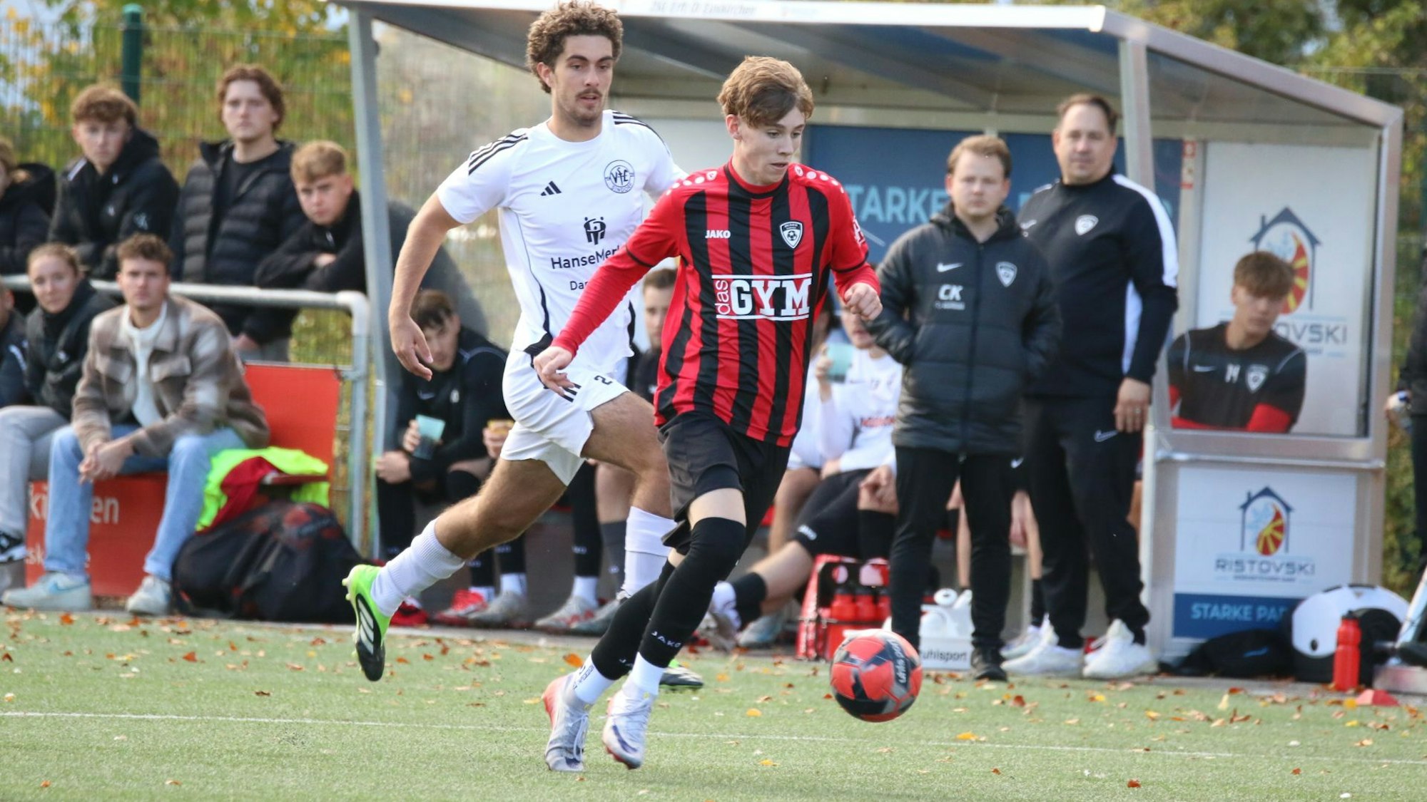 Aaron Mertens läuft seinem Gegenspieler davon. Im Hintergrund schauen Trainer, Betreuer und Mitspieler gespannt zu.