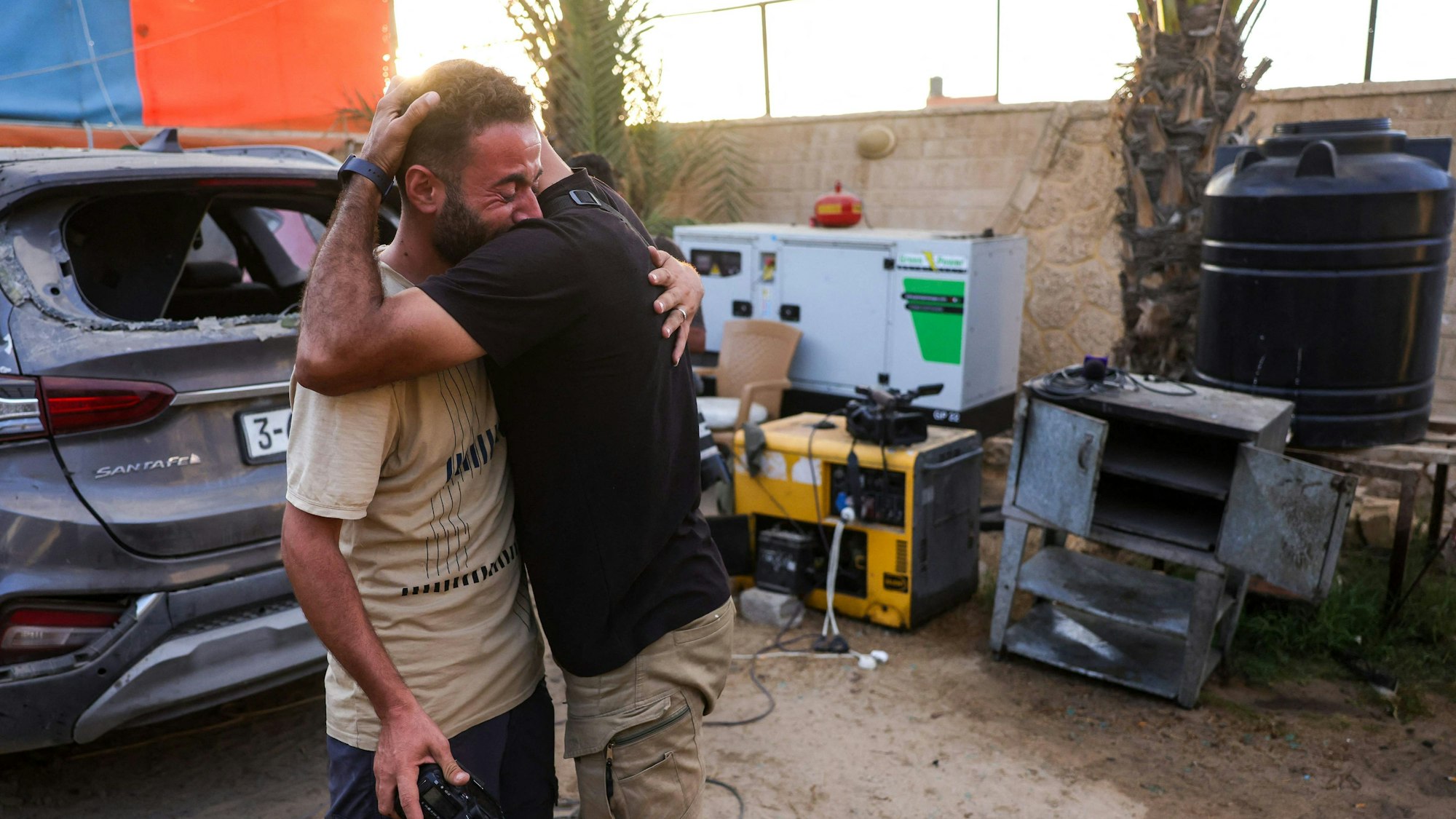 Zwei Menschen umarmen sich. Im Hintergrund ein Fahrzeug mit zerstörter Heckscheibe.