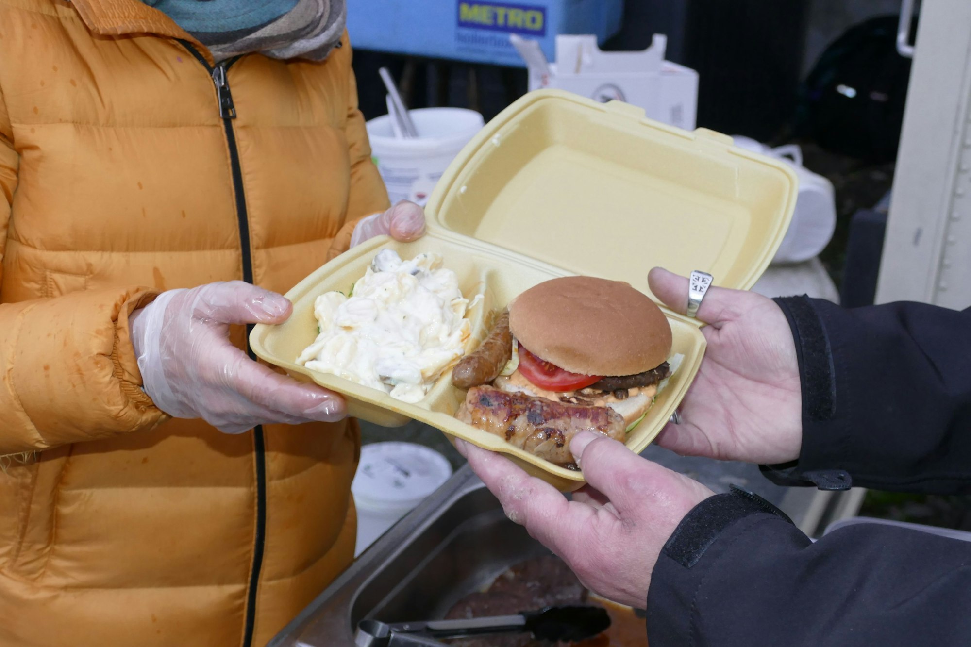 Zwei Händepaare halten eine Styropor-Schale mit Hamburger und Kartoffelsalat.