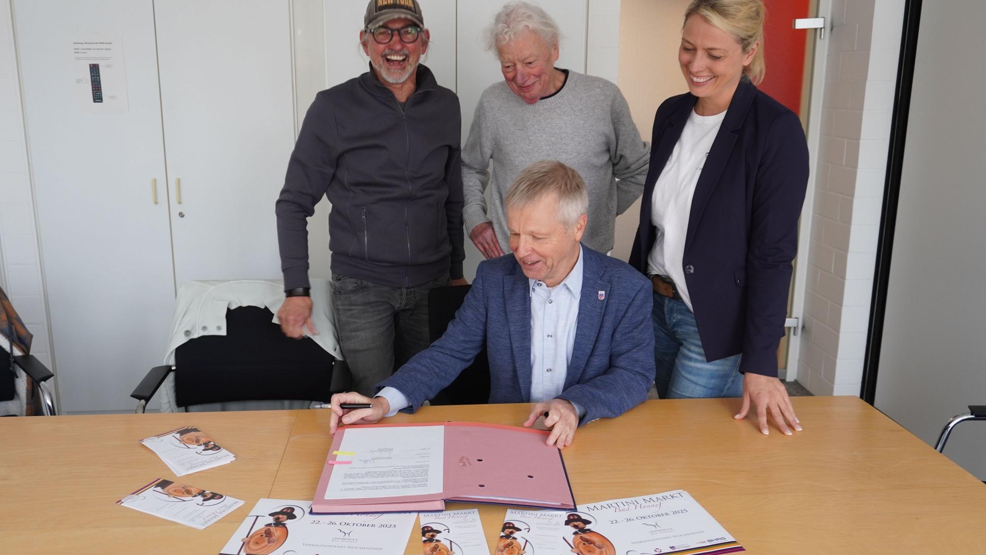 Drei Männer und eine Frau. Ein Mann sitzt an einem Tisch vor einer Dokumentenmappe.