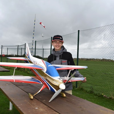 Marian Friedrich steht hinter seinem Modellflugzeug, ein weißer Doppeldecker mit blauen, roten und goldenen Akzenten.