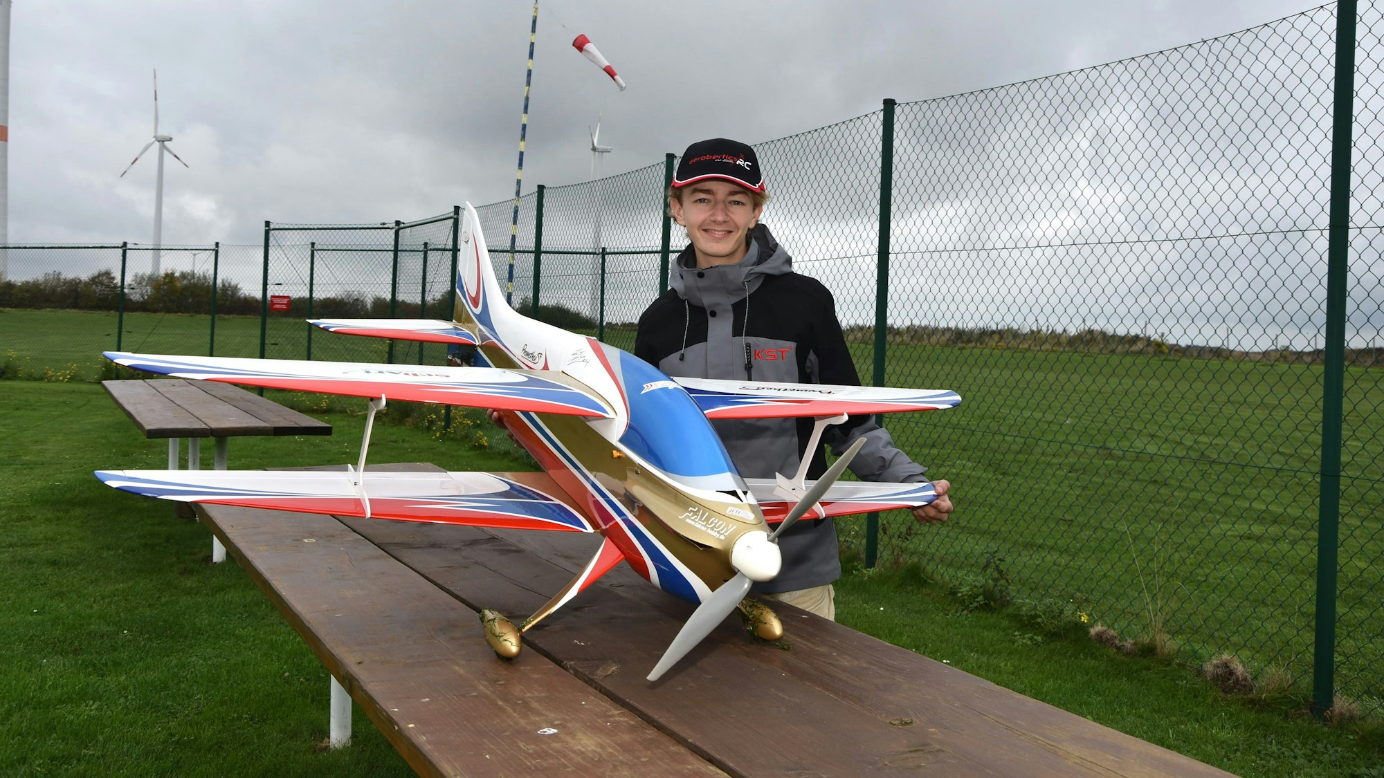 Marian Friedrich steht hinter seinem Modellflugzeug, ein weißer Doppeldecker mit blauen, roten und goldenen Akzenten.