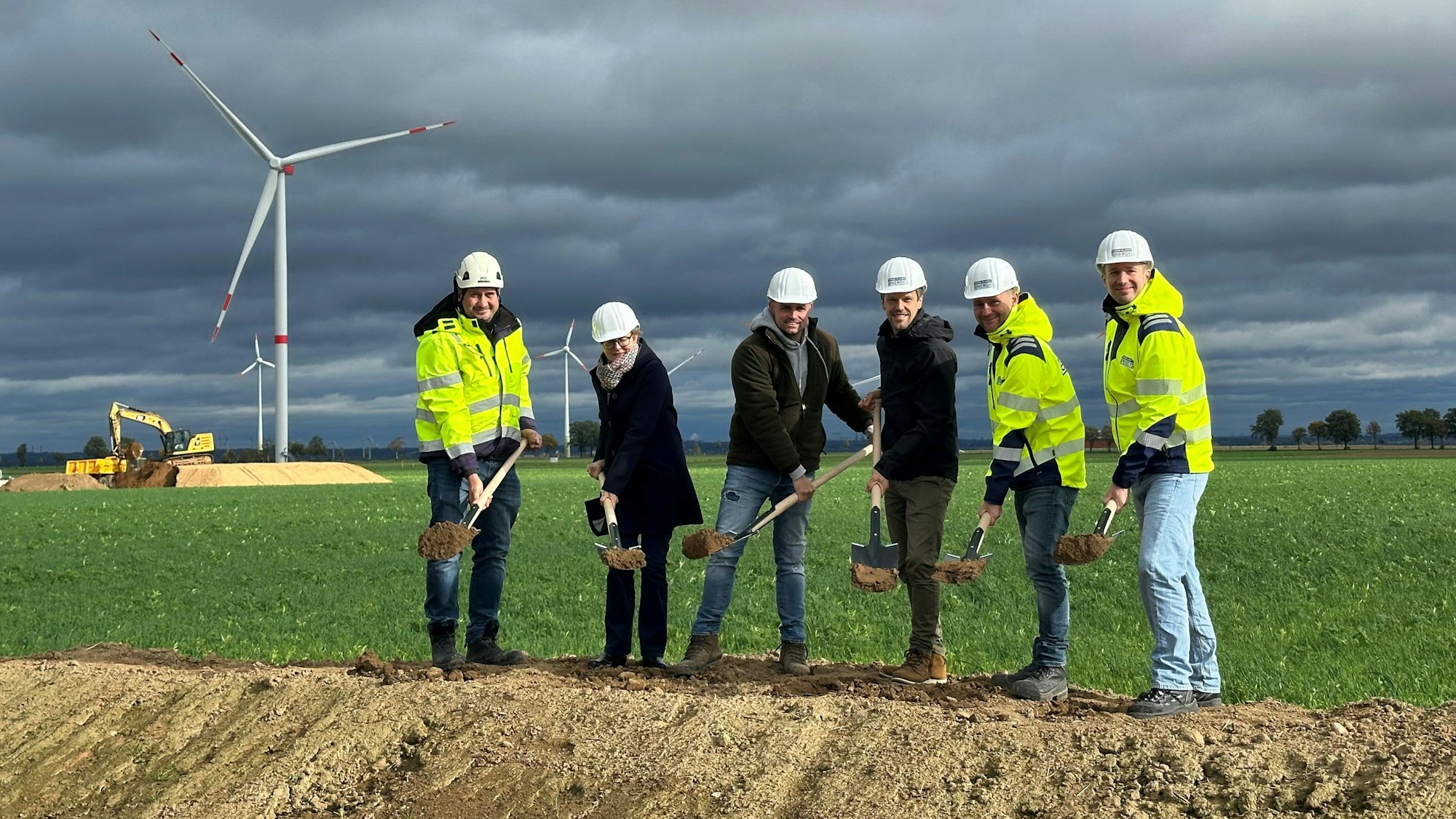 Männer und eine Frau mit Helmen halten Spaten mit Erde.