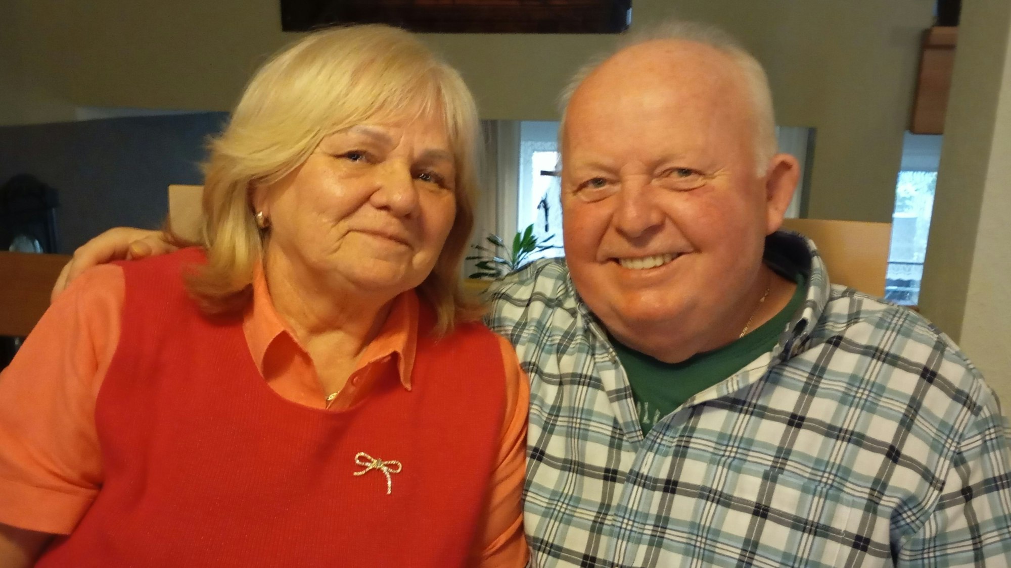 Maria Sander trägt einen roten Pollunder und ein rötliches Hemd. Georg Sander trägt ein kariertes Hemd. Er hat den Arm um seine Frau gelegt.