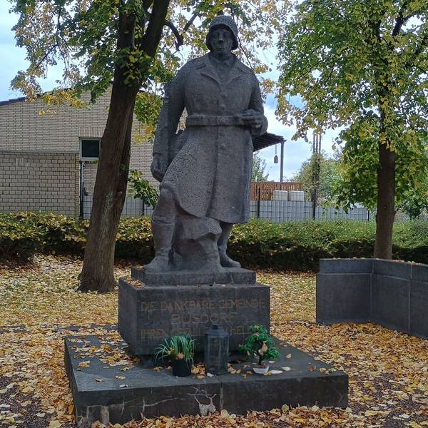 Zu sehen ist ein Steinsoldat mit Helm und Uniform. Auf seinem Podest steht: „Die dankbare Gemeinde Büsdorf ihren gefallenen Helden.“