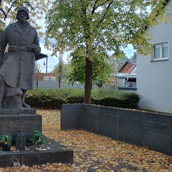 Zu sehen ist ein Steinsoldat mit Helm und Uniform. Auf seinem Podest steht: „Die dankbare Gemeinde Büsdorf ihren gefallenen Helden.“