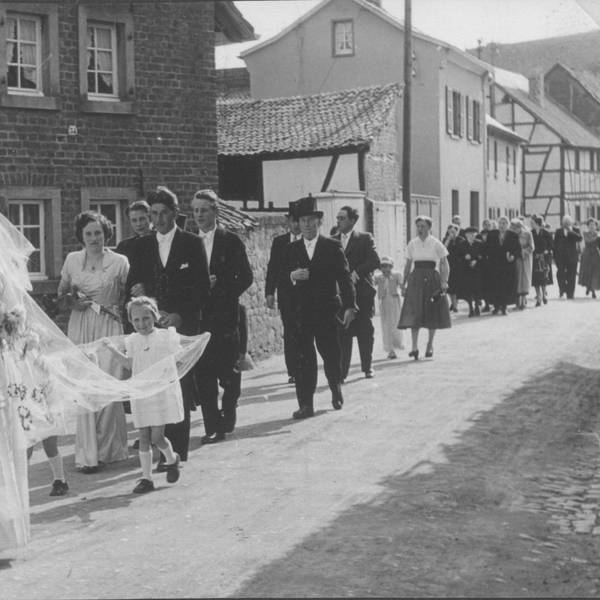 Das Bild zeigt eine Hochzeitsgesellschaft, die eine Dorfstraße entlanggeht.