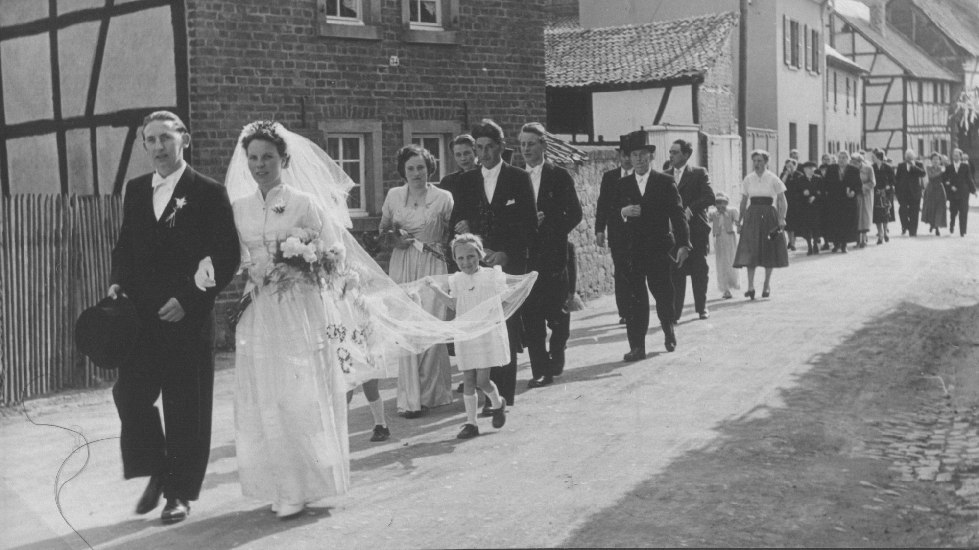 Das Bild zeigt eine Hochzeitsgesellschaft, die eine Dorfstraße entlanggeht.